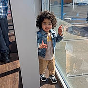 Emanveer is registered to the contest to win money with this photo: child, toddler, curly_hair, denim_jacket, striped_shirt, khaki_pants, sneakers, reflection, glass_window, pillar, indoor, sunlight, wooden_floor, playful, person, casual_clothing, daylight, smile, standing, pose