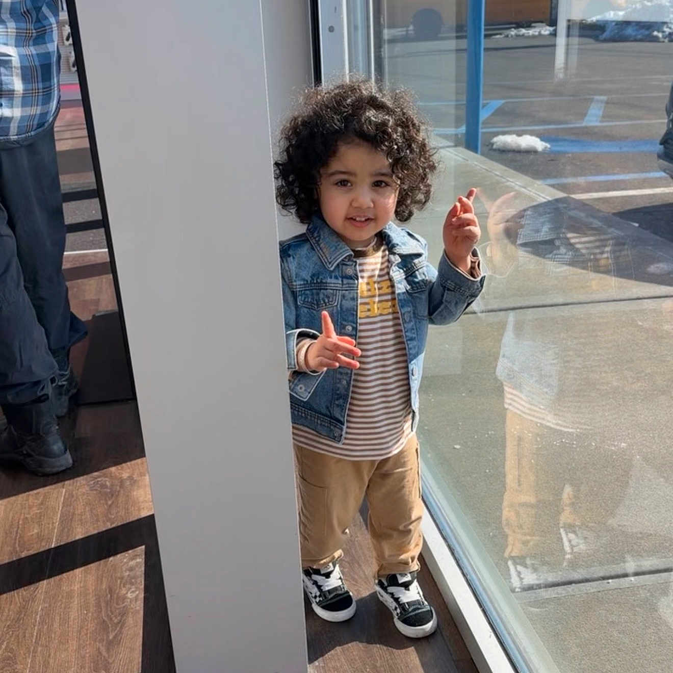 Emanveer is registered to the contest to win money with this photo: child, toddler, curly_hair, denim_jacket, striped_shirt, khaki_pants, sneakers, reflection, glass_window, pillar, indoor, sunlight, wooden_floor, playful, person, casual_clothing, daylight, smile, standing, pose