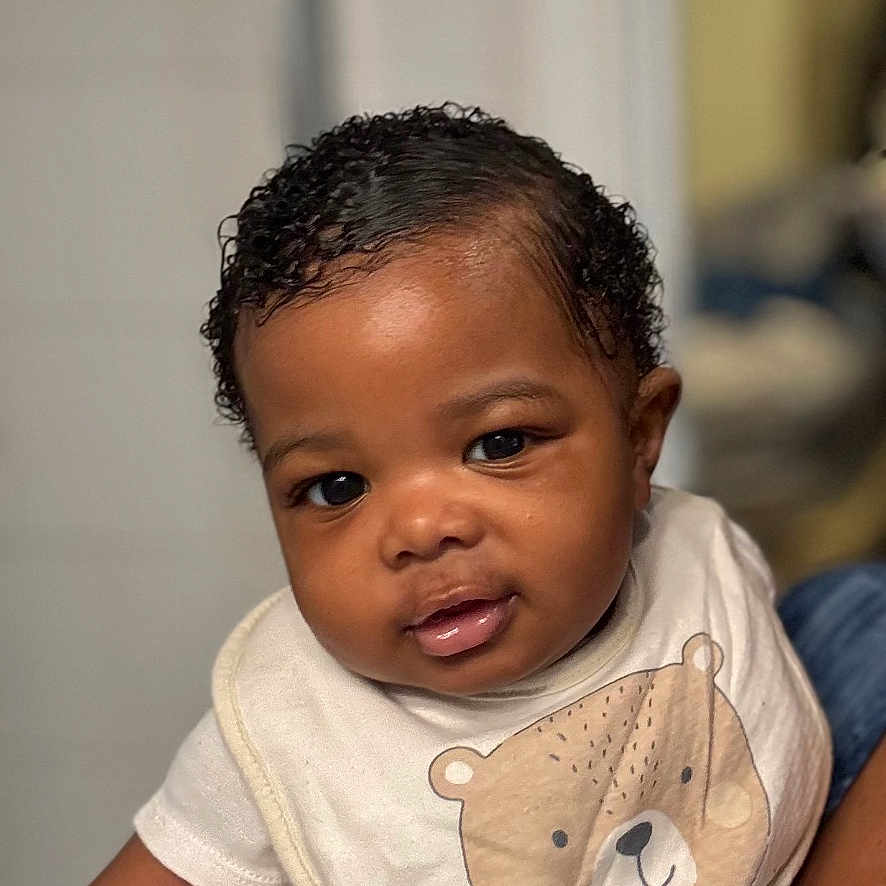 Prestyn is registered to the contest to win money with this photo: adorable, baby, bear_print, bib, child, closeup, clothing, curly_hair, cute, face, hand, holding, human, indoor, infant, person, portrait, skin, smile, young_child