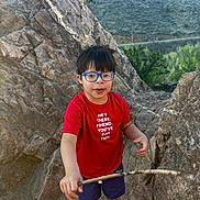 Xavier joined the competition — help win amazing prizes! child, boy, glasses, red_shirt, stick, rocks, mountains, outdoor, nature, hiking, smiling, shorts, black_shoes, daylight, adventure, landscape, person, casual_clothing, greenery, rocky_terrain