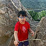 child, boy, glasses, red_shirt, stick, rocks, mountains, outdoor, nature, hiking, smiling, shorts, black_shoes, daylight, adventure, landscape, person, casual_clothing, greenery, rocky_terrain