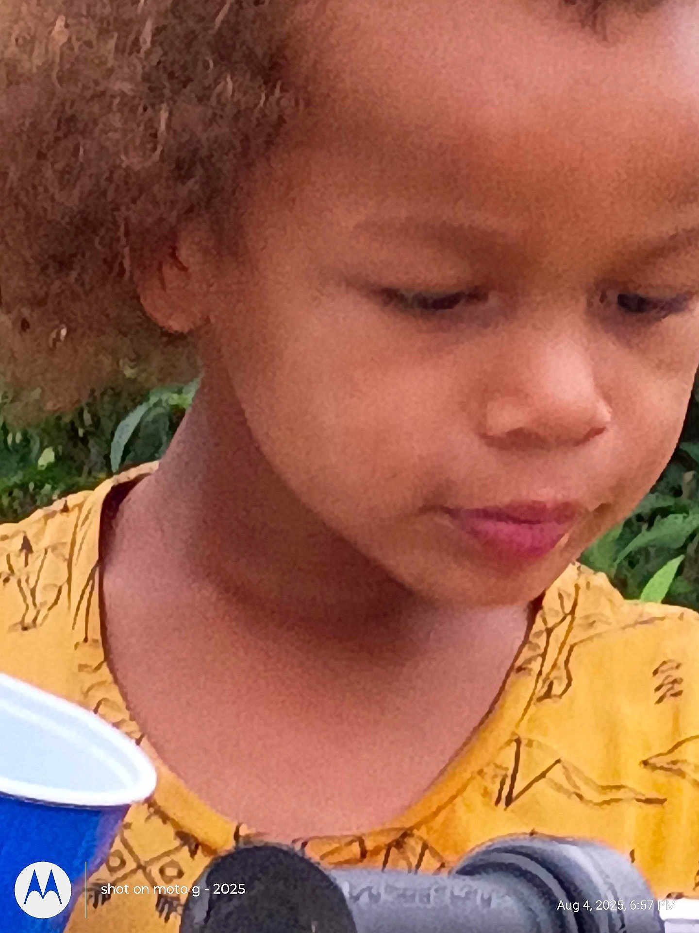 Kari is registered to the contest to win money with this photo: child, curly_hair, yellow_shirt, close_up, outdoor, greenery, cup, thoughtful, person, face, portrait, skin, young, expression, casual, daylight, nature, head, clothing, focus