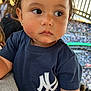 toddler, child, face, person, clothing, tshirt, stadium, crowd, sports, event, earrings, hair, expression, side_view, young_child, indoor, lighting, portrait, casual, spectators