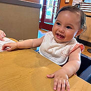 Raiden is registered to the contest to win money with this photo: baby, child, smile, happy, table, bib, indoor, restaurant, wooden_table, face, person, cute, toddler, skin, hair, arm, hand, seat, light, window
