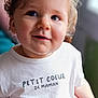 Leyann participe au concours pour gagner de l'argent avec cette photo : child, toddler, curly_hair, blue_eyes, white_shirt, text_on_clothing, indoor, portrait, smiling, cute, baby, person, soft_light, closeup, cute_expression, happy, hand, casual_clothing, human, infant