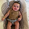 baby, child, infant, smiling, portrait, sitting, bouncer, baby_chair, harness, patterned_fabric, indoor, home, brown_outfit, hair_bow, ponytail, bracelets, socks, happy, face, furniture