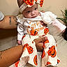 baby, child, infant, portrait, halloween, pumpkin, costume, bow, headband, white_clothing, orange, jack_o_lantern, sitting, held, adult_arms, indoor, plant, couch, facial_expression, photography