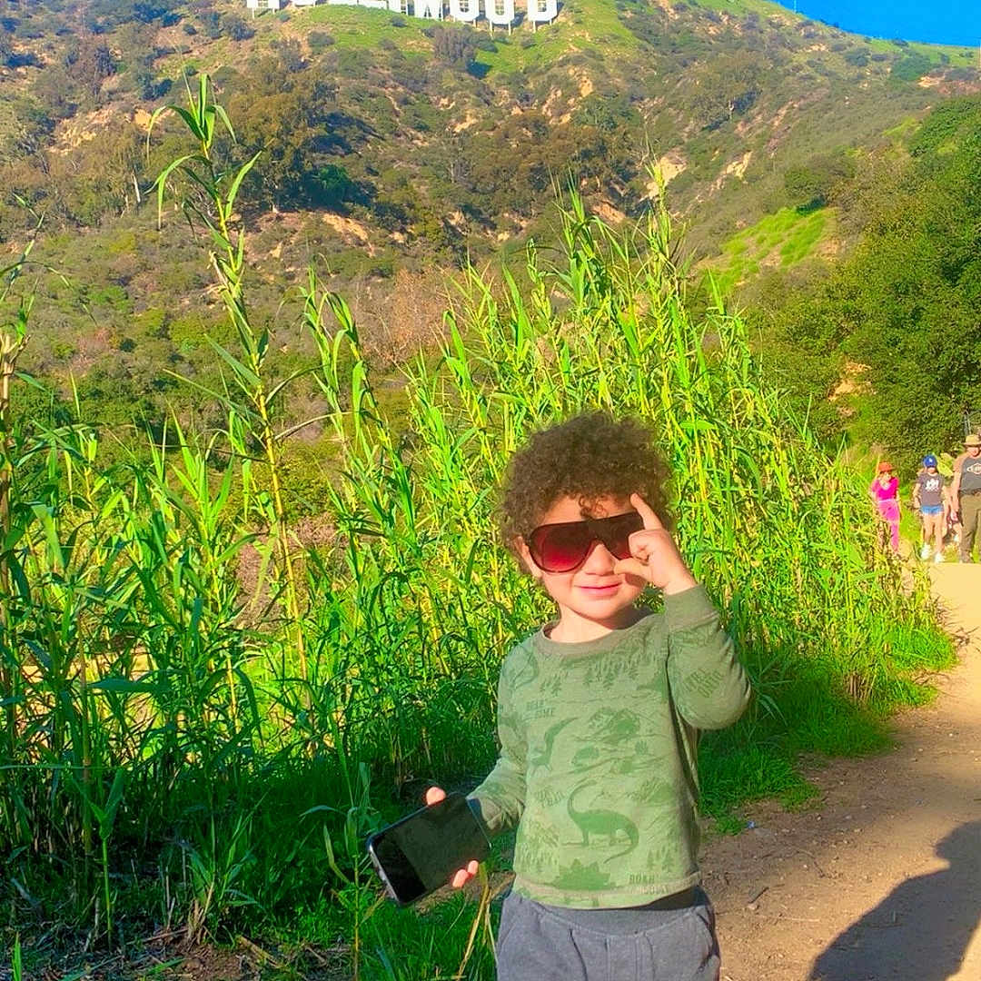 Shahem joined the competition — help win amazing prizes! boy, casual_clothing, child, curly_hair, daytime, gray_pants, green_shirt, hill, hollywood_sign, nature, outdoor, people_in_background, playful, red_shoes, shadow, smartphone, sunglasses, sunny, trail, vegetation