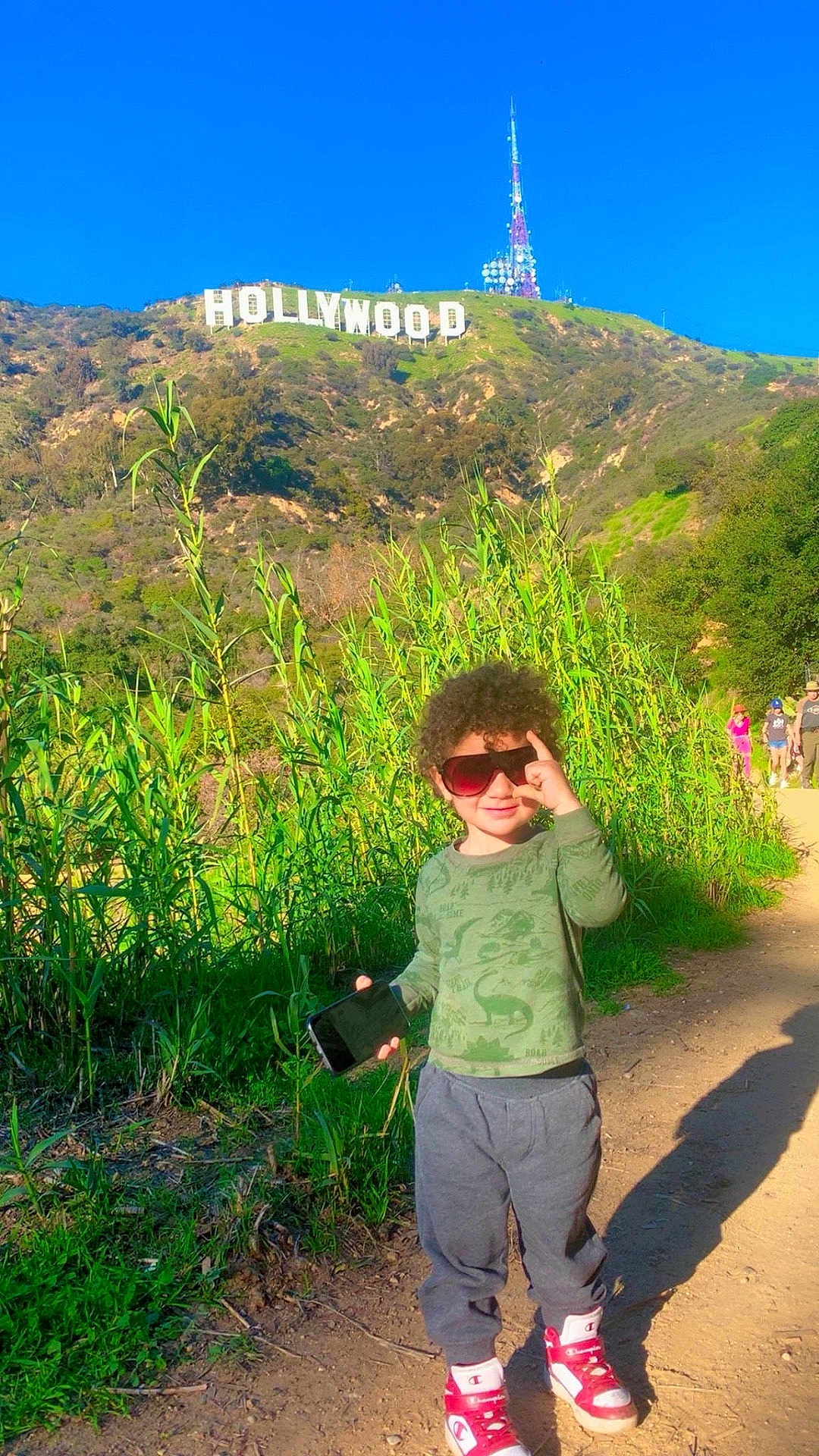 Shahem joined the competition — help win amazing prizes! child, boy, curly_hair, sunglasses, smartphone, green_shirt, gray_pants, red_shoes, outdoor, trail, hollywood_sign, hill, vegetation, sunny, daytime, shadow, nature, people_in_background, casual_clothing, playful