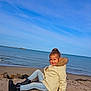 child, girl, smile, coat, fur_hood, jeans, boots, beach, sea, sky, rocks, sitting, outdoor, portrait, sunny, relaxed, casual, happy, nature, daytime