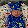baby, infant, swaddled, blue_clothing, hat, dinosaur_pattern, pillow, colorful, animal_print, rainbow, expression, grumpy, cute, face, child, portrait, indoors, soft_fabric, newborn, lying_down