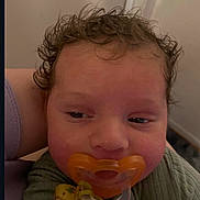 Rome is registered to the contest to win money with this photo: baby, pacifier, curly_hair, sleepy, close_up, infant, child, person, green_clothing, hand, resting, indoors, soft_lighting, skin, face, mouth, cute, young, pacifier_clip, comfort