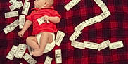 Leo joined the competition — help win amazing prizes! baby, infant, red_shirt, diaper, blanket, plaid, money, fake_money, question_mark, curious, lying_down, indoor, child, face, head, hand, foot, fabric, pattern, currency