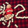 baby, infant, red_shirt, diaper, blanket, plaid, money, fake_money, question_mark, curious, lying_down, indoor, child, face, head, hand, foot, fabric, pattern, currency