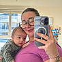 woman, baby, infant, mirror_selfie, smartphone, glasses, purple_shirt, tattoo, ring, bracelet, window, indoor, family, holding, portrait, reflection, newborn, cozy, home, smile