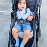 toddler, child, stroller, blue_socks, plaid_shirt, shorts, outdoor, curly_hair, urban, sidewalk, safety_harness, baby, person, casual_clothing, cute, young_child, daylight, portrait, seated, expression