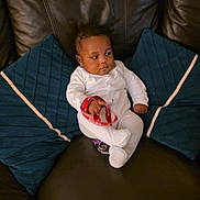 Hugh is registered to the contest to win money with this photo: baby, infant, couch, leather, blue_cushions, white_onesie, wristband, indoor, child, sitting, furniture, relaxed, looking_away, hand, footwear, person, home, comfort, portrait, cute