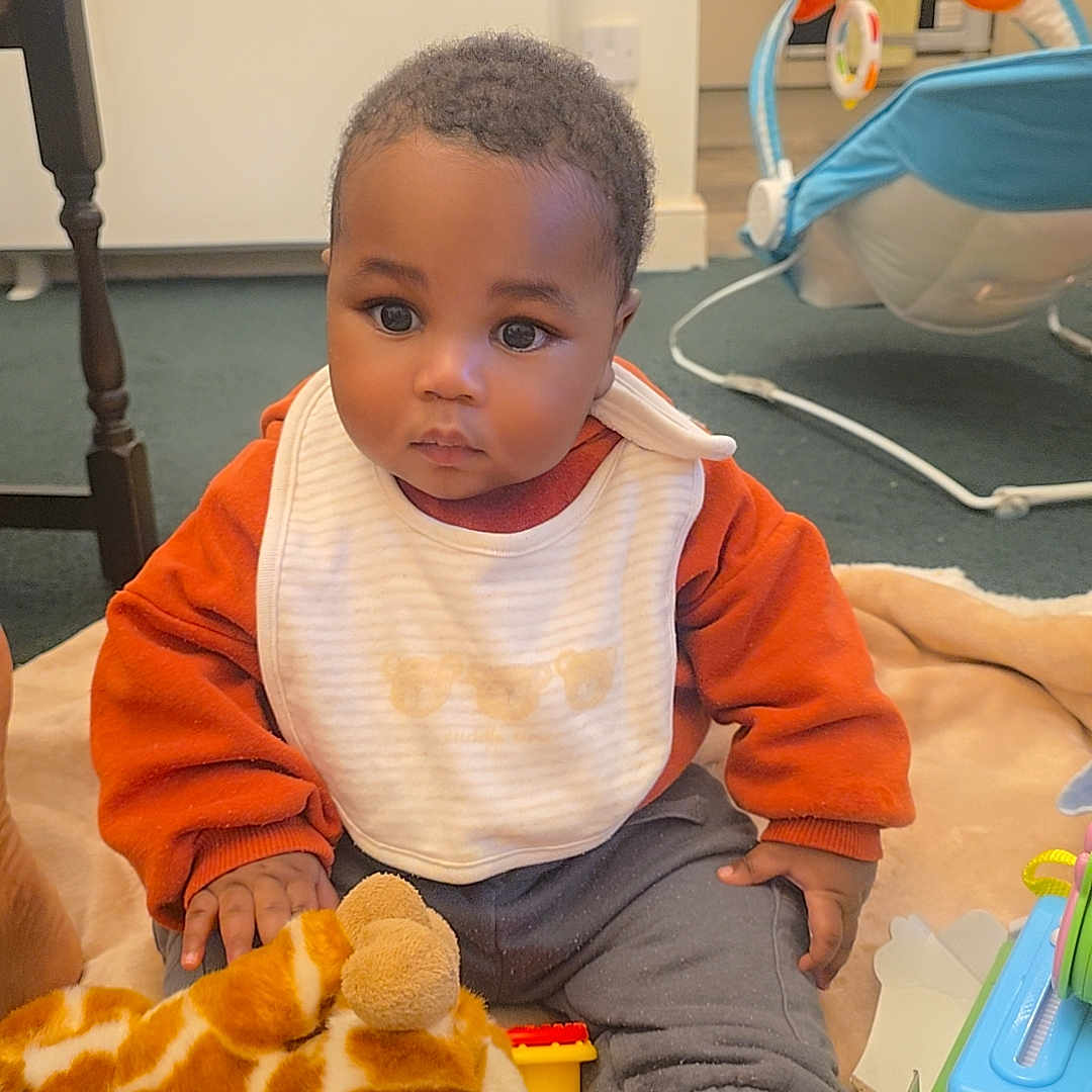 Hugh is registered to the contest to win money with this photo: baby, bib, blanket, carpet, child, clothing, curious, floor, footwear, furniture, indoor, infant, kitchen, person, playtime, plush_giraffe, polka_dot, sitting, soft_toy, toy