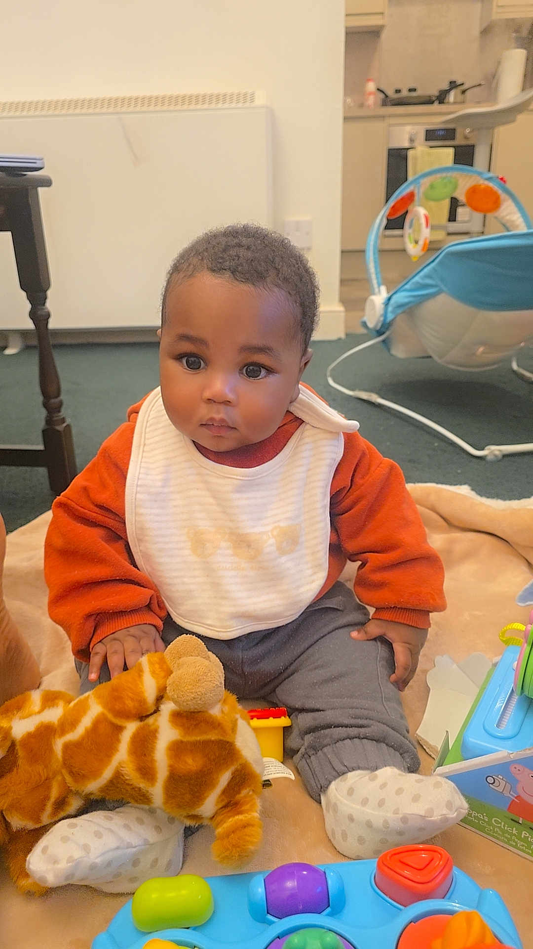 Hugh is registered to the contest to win money with this photo: baby, child, indoor, toy, plush_giraffe, blanket, bib, sitting, curious, infant, clothing, playtime, floor, carpet, person, footwear, polka_dot, kitchen, furniture, soft_toy