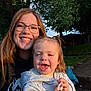 woman, child, toddler, smile, glasses, outdoor, sunlight, happy, face, pacifier, hoodie, grass, trees, pavement, backyard, daylight, person, nature, family, casual