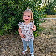 Oaklynn is registered to the contest to win money with this photo: toddler, child, smile, outdoor, grass, greenery, bush, sunlight, happy, summer, nature, sandals, leggings, floral_top, hair_bow, person, standing, daylight, cute, portrait