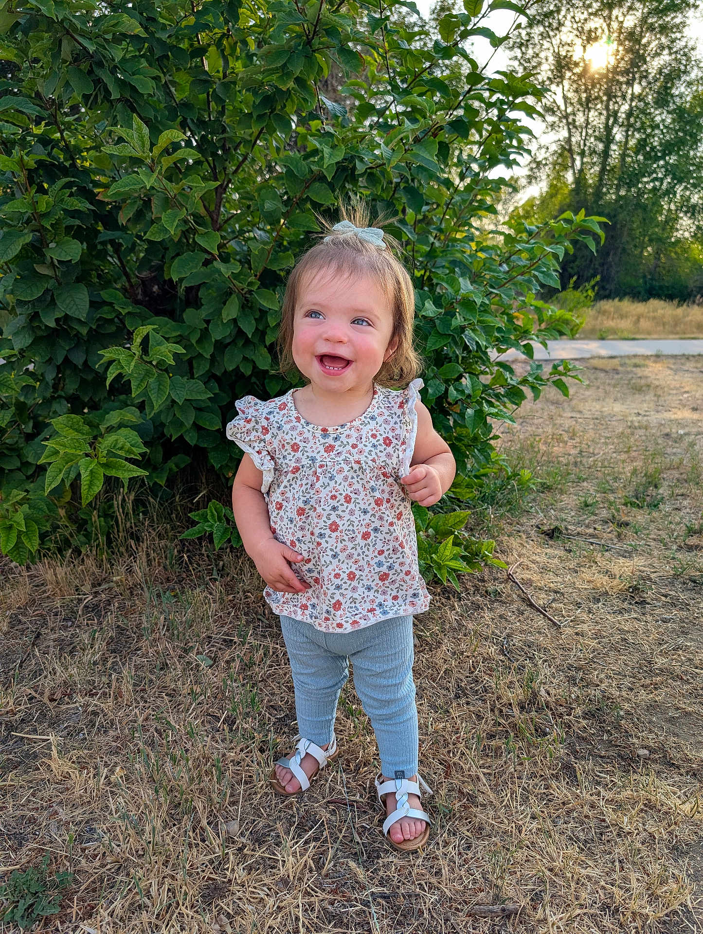 Oaklynn is registered to the contest to win money with this photo: toddler, child, smile, outdoor, grass, greenery, bush, sunlight, happy, summer, nature, sandals, leggings, floral_top, hair_bow, person, standing, daylight, cute, portrait