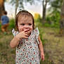 toddler, child, outdoor, nature, trees, grass, blurred_background, floral_top, blue_pants, smiling, cute, person, young_child, playful, sunlight, greenery, portrait, casual_clothing, happy, finger_in_mouth