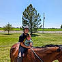 adult, child, horse, helmet, glasses, grass, tree, fence, rope, outdoor, sunny, field, riding, nature, smile, person, animal, daylight, leisure, family