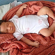 Rain is registered to the contest to win money with this photo: baby, sleeping, blanket, onesie, infant, resting, cozy, soft_texture, pink, white_clothing, curly_hair, peaceful, portrait, indoor, child, cute, relaxation, newborn, comfort, toddler