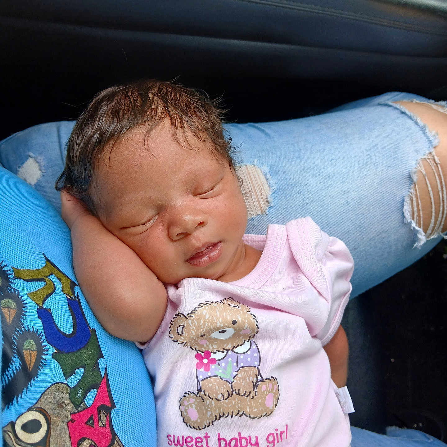 Rain is registered to the contest to win money with this photo: baby, sleeping, infant, pink_onesie, teddy_bear, text_on_clothing, person, lap, ripped_jeans, blue_clothing, car_interior, door_controls, cute, peaceful, child, portrait, human, resting, casual_wear, young_child