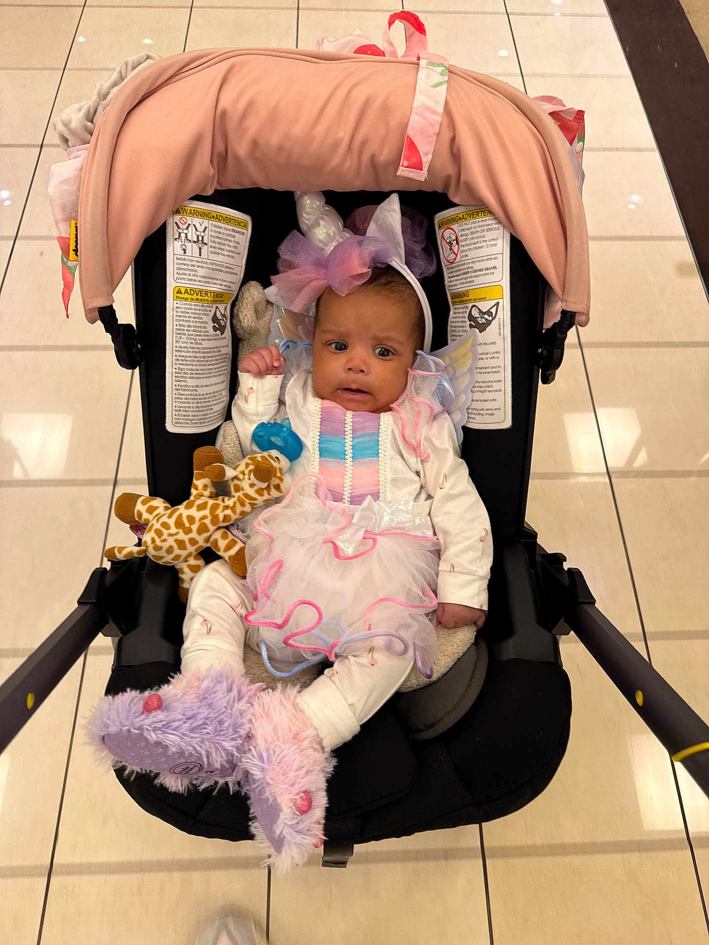 Kali J joined the competition — help win amazing prizes! baby, stroller, unicorn_costume, plush_toy, giraffe, slippers, floor_tiles, headband, wings, child, indoor, cute, infant, clothing, pink, purple, white, person, portrait, toy