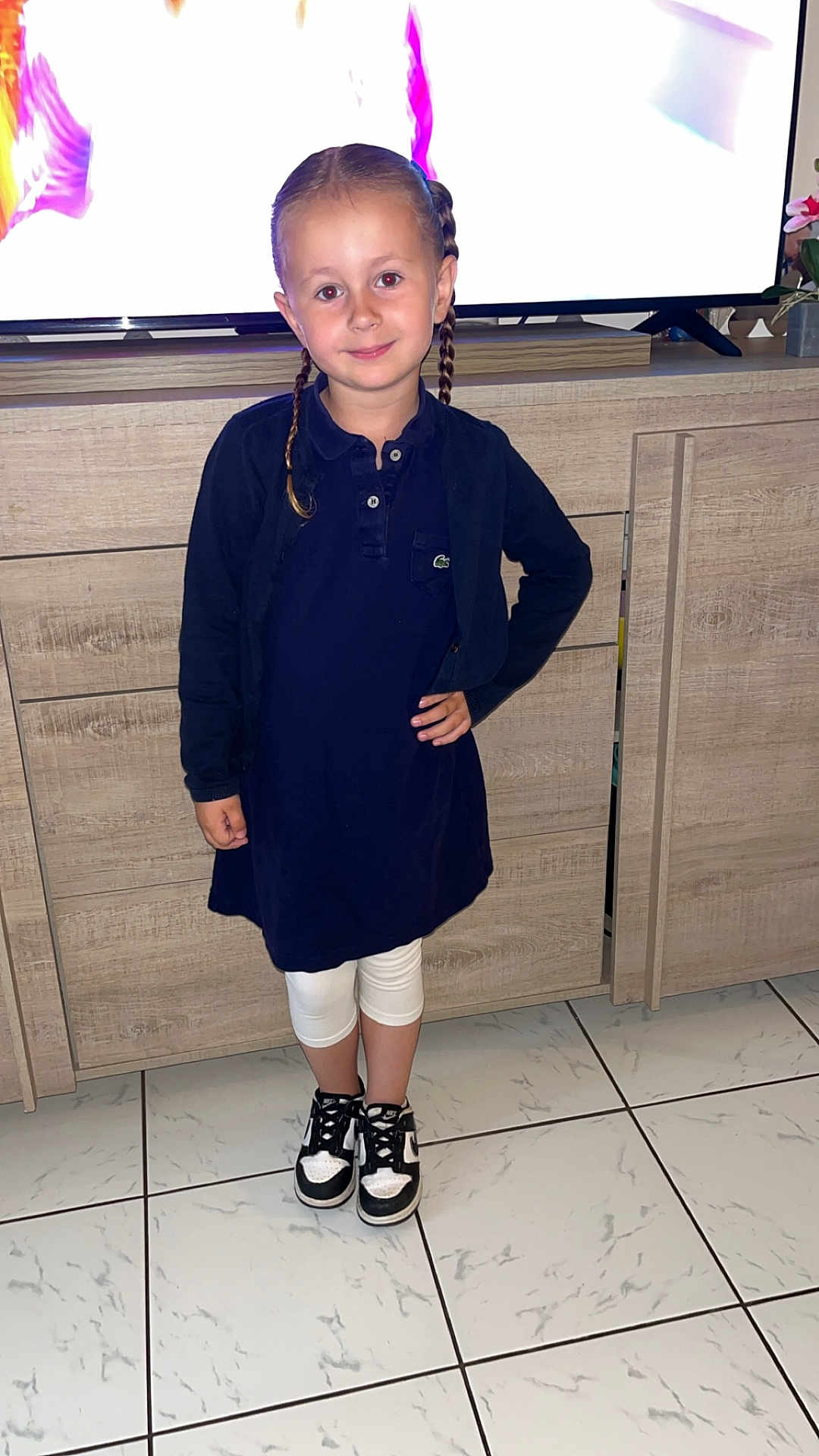 Maëlyne a rejoint le concours — aidez-le/la à gagner de superbes lots ! child, girl, braids, navy_dress, white_leggings, sneakers, indoor, tiled_floor, wooden_cabinet, television, smiling, standing, pose, floor, clothing, person, hair, footwear, casual, home