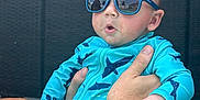 Carson is registered to the contest to win money with this photo: baby, child, sunglasses, tattoo, swimsuit, blue_clothing, outdoor, person, arm, hand, face, expression, poolside, concrete, summer, sunny, reflection, cute, infant, holding
