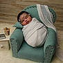 Riot is registered to the contest to win money with this photo: baby, swaddled, sleeping, armchair, teal, pearl_headband, pillow, blanket, side_table, iced_coffee, laptop, rug, wooden_floor, wooden_wall, infant, cozy, indoor, cute, peaceful, portrait