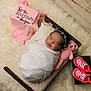 baby, infant, sleeping, swaddled, blanket, wooden_bed, crocheted_toy, pink_teddy_bear, baby_shoes, pink_sneakers, onesie, text_on_clothing, fur_rug, headband, face, newborn, cozy, cute, indoor, portrait