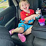 child, smiling, car_seat, toy, blue_shorts, red_shirt, pink_shoes, car_interior, seatbelt, cup, straw, happy, playful, window, daylight, vehicle, seat, toy_truck, young_child, casual_clothing