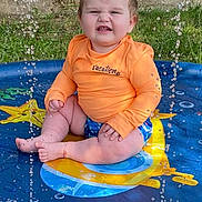 Oliver is registered to the contest to win money with this photo: baby, child, splash_pool, water, orange_shirt, blue_shorts, summer, outdoor, grass, brick_wall, window, sitting, playful, water_droplets, face, expression, fun, sunny, cute, vacation