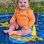 baby, child, splash_pool, water, orange_shirt, blue_shorts, summer, outdoor, grass, brick_wall, window, sitting, playful, water_droplets, face, expression, fun, sunny, cute, vacation