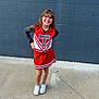 Madeline is registered to the contest to win money with this photo: child, cheerleader, uniform, red, white, smile, pose, concrete, wall, outdoor, young, girl, hands_on_hips, long_hair, casual, shoes, sport, cheer, portrait, confident