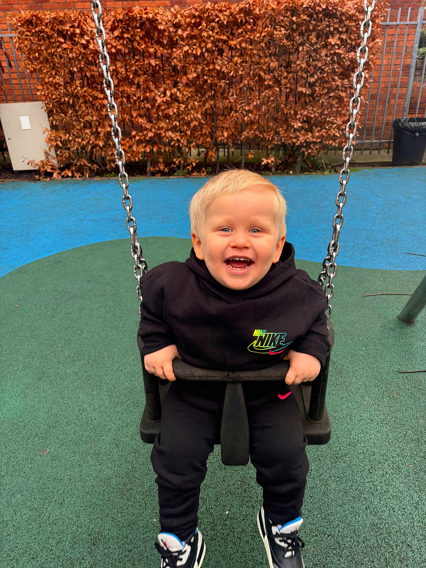 Reggie is registered to the contest to win money with this photo: child, toddler, swing, playground, laughing, happy, black_clothing, nike, hoodie, sneakers, blonde_hair, outdoor, fence, hedge, rubber_surface, green, blue, metal_chain, seat, grip