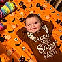 baby, black_cat, brown_clothing, child, colorful_toy, crib, cute, face, halloween_theme, happy, indoor, infant, lying_down, onesie, orange_sheet, person, pumpkin, skull, smile, toy