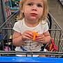 Aarika is registered to the contest to win money with this photo: toddler, child, shopping_cart, snack, cheese_puffs, store, supermarket, aisle, blond_hair, messy_hair, blue_eyes, white_shirt, sitting, face, food, casual, indoor, people, retail, curious