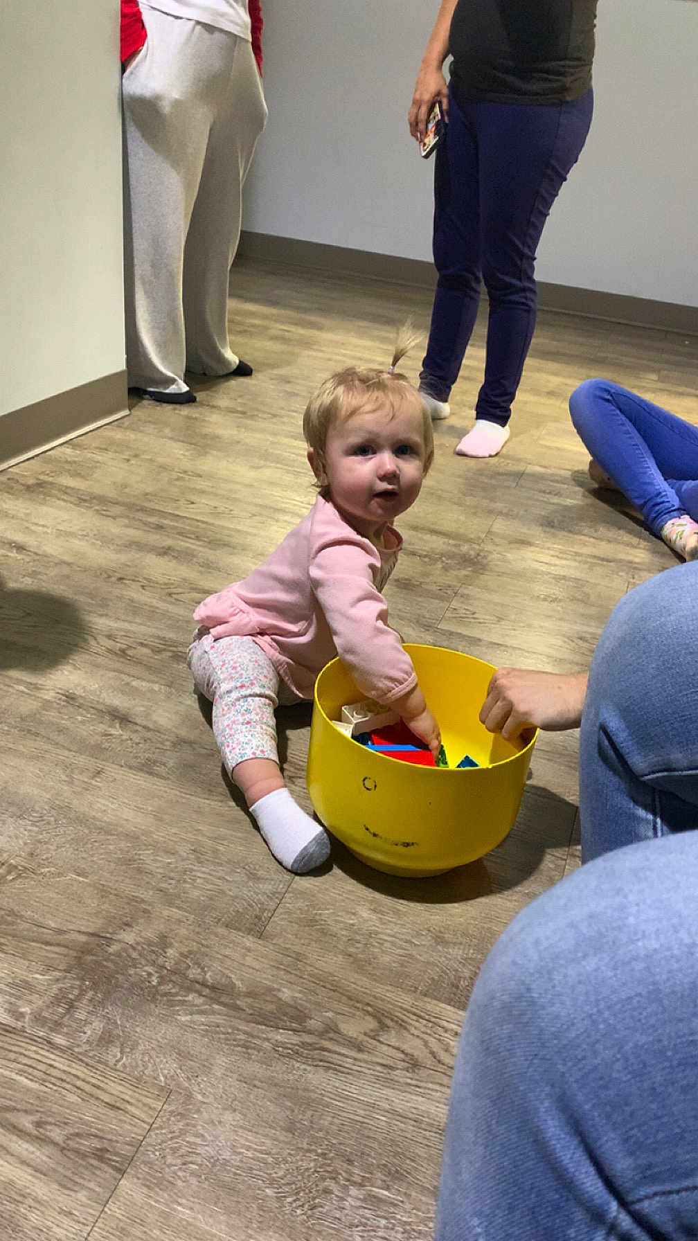 Adaline is registered to the contest to win money with this photo: toddler, child, toy, yellow_bucket, indoor, wooden_floor, person, pants, sock, sitting, playing, casual, people, light, hair, floor, hand, clothing, room, curious