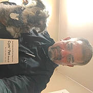 Maxx is registered to the contest to win money with this photo: beard, black_shirt, box, ceiling, collar, cute, dog, fur, holding, human, indoor, light, looking_at_camera, man, pet, portrait, short_hair, smiling, tag, wall