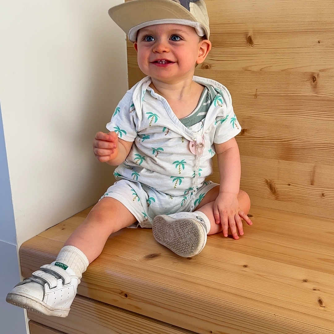 Elio a rejoint le concours — aidez-le/la à gagner de superbes lots ! baby, blue_eyes, cap, casual_clothing, child, cute, happy, hat, indoor, light_skin, palm_tree_pattern, playful, shorts, sitting, smiling, socks, toddler, white_shoes, wood_paneling, wooden_bench