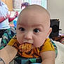baby, blue_eyes, blurred_background, child, closeup, clothing, curious, expression, face, food, furniture, hairless, head, highchair, indoor, person, reaching, snack, television, yellow_shirt