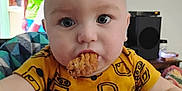 Landen is registered to the contest to win money with this photo: baby, child, face, blue_eyes, high_chair, cookie, snack, yellow_shirt, patterned_clothing, indoor, television, person, woman, blurred_background, furniture, room, table, head, close_up, curious