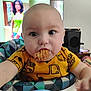 baby, blue_eyes, blurred_background, child, close_up, cookie, curious, face, furniture, head, high_chair, indoor, patterned_clothing, person, room, snack, table, television, woman, yellow_shirt
