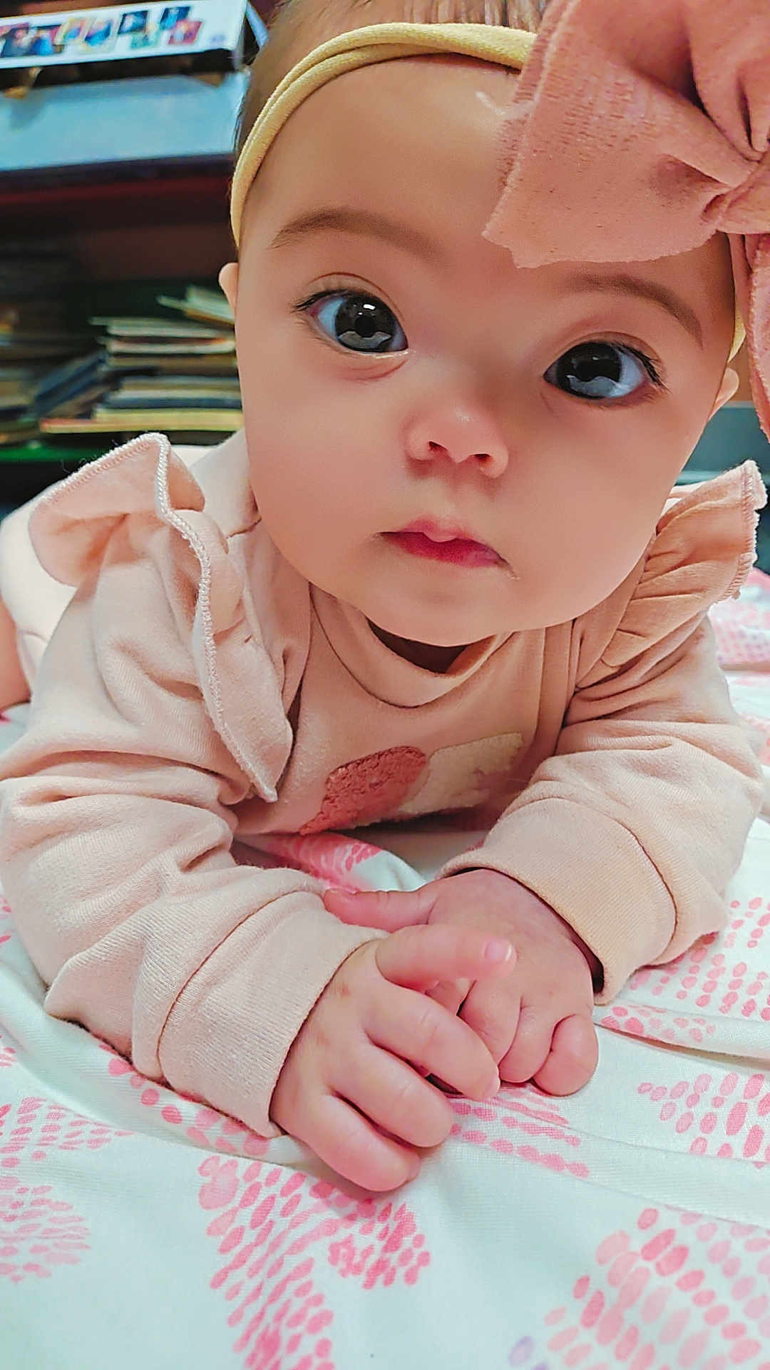Oliviah is registered to the contest to win money with this photo: baby, infant, close_up, pink_clothing, headband, bow, hands, blanket, indoor, curious_eyes, soft_fabric, child, portrait, cute, face, young_child, laying_down, patterned_blanket, warm_lighting, bookshelf
