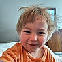 child, toddler, smile, face, hair, orange_shirt, indoor, bed, furniture, person, cute, happy, portrait, casual, young_child, messy_hair, room, cozy, closeup, home